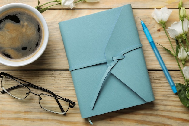 Beautiful blue notebook, cup of coffee, glasses, and white flowers on a natural wooden table. Top view