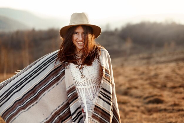 Foto bella boho donna hipster sorridente e divertirsi al tramonto in montagna indossando cappello e poncho