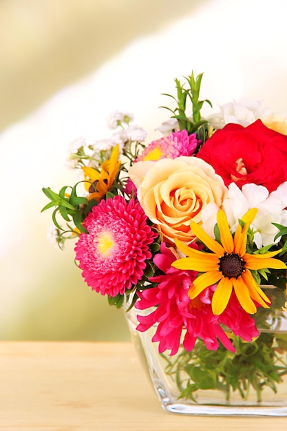 Beautiful bouquet of bright flowers in glass vase on wooden table on bright background