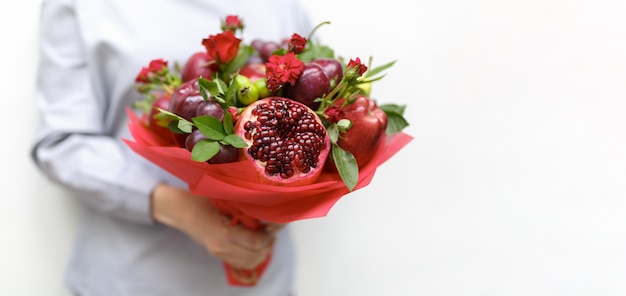 Beautiful bouquet consisting of pomegranate, apples, grapes, plums and scarlet roses in the hands of a woman on a white as a blank for a greeting card