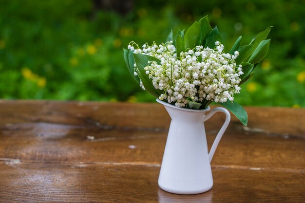 庭の木製テーブルの上の白いエナメルの花瓶にスズランの美しい花束、クローズアップ