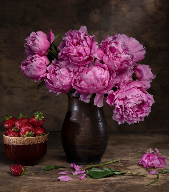 Beautiful bouquet of pink peonies in vase and strawberries in a bowl on a wooden table