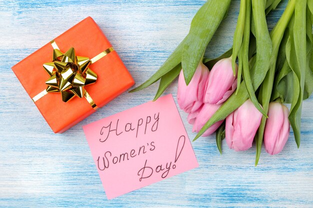 Beautiful bouquet of pink tulips flowers, gift box and text Happy Women's Day on paper on blue wooden surface