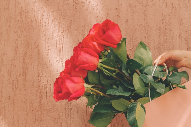 粗い壁に対して女性の手でパステル調のピンクのパッケージにバラの美しい花束