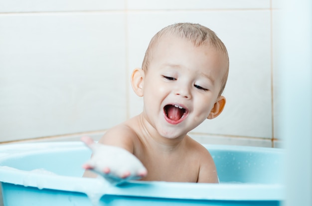 Красивый мальчик купает малыша в ванной чистой и гигиеничной, смотрит на руку, которая в пене