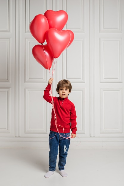 Beautiful boy in jeans and sweater holding heart balloons on white background