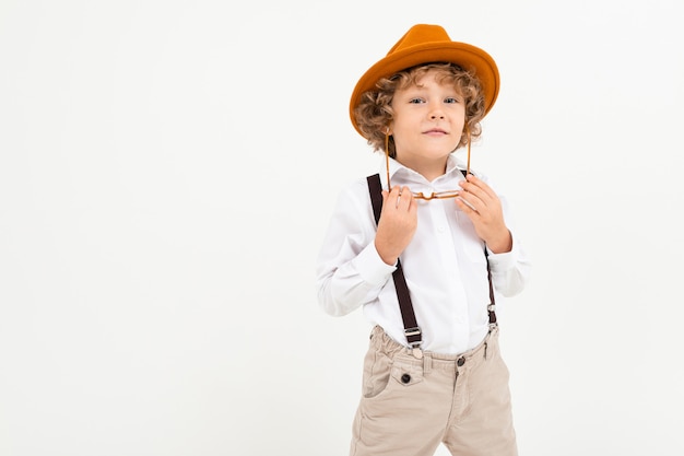 Beautiful boy with curly hair in white shirt, brown hat, glasses with black suspenders stands isolated on white