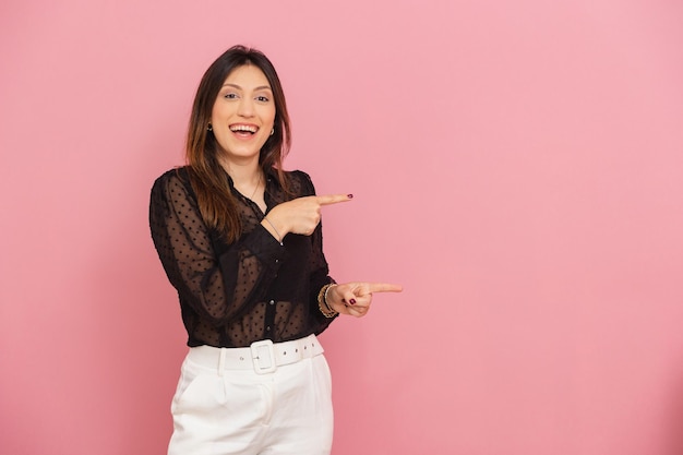 아름다운 브라질 백인 여성 핑크색 배경은 그녀의 광고 네거티브 공간 옆에 손가락으로 제품이나 광고를 보여주며 수용적인 미소를 짓고 있습니다.