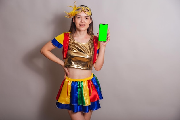 Beautiful brazilian caucasian woman wearing frevo carnival
clothes wearing a mask holding smartphone with green screen in
chroma