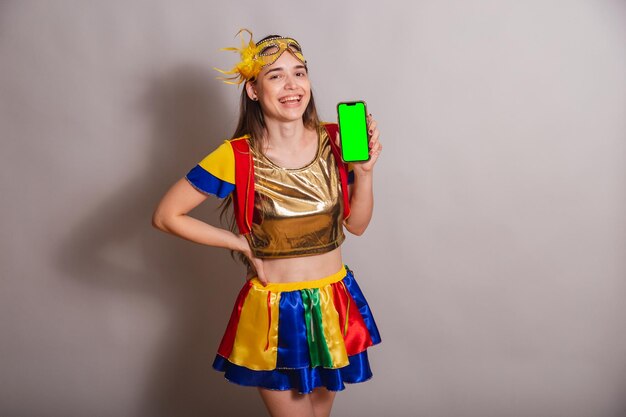 Beautiful brazilian caucasian woman wearing frevo carnival\
clothes wearing a mask holding smartphone with green screen in\
chroma