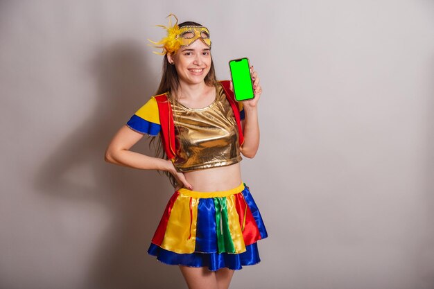 Beautiful brazilian caucasian woman wearing frevo carnival\
clothes wearing a mask holding smartphone with green screen in\
chroma