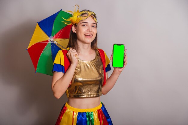 Beautiful brazilian caucasian woman wearing frevo carnival\
clothes wearing a mask holding smartphone with green screen in\
chroma