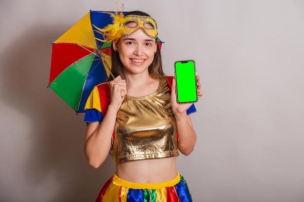 Beautiful brazilian caucasian woman wearing frevo carnival\
clothes wearing a mask holding smartphone with green screen in\
chroma