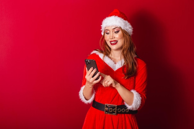 Beautiful Brazilian redhaired woman dressed in Christmas clothes Santa Claus holding smartphone typing