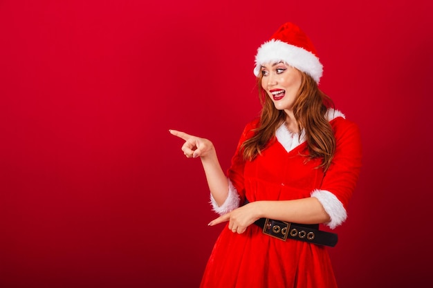 クリスマスの服を着た美しいブラジルの赤髪の女性サンタ クロースが左に何かを示す
