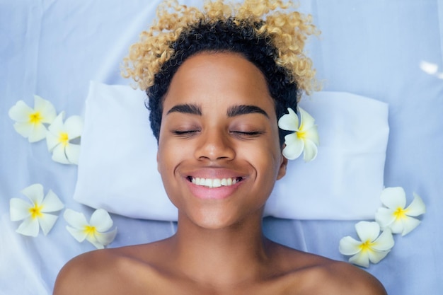 Beautiful brazilian smiling girl lying on table with towel and white flowers massaging and pampering herself in luxury resort spa centre in Bali