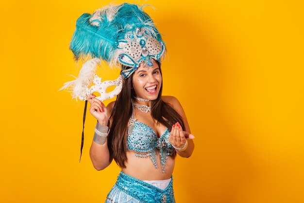 Beautiful brazilian woman in blue and white carnival clothes with crown of feathers and mask inviting to the carnival