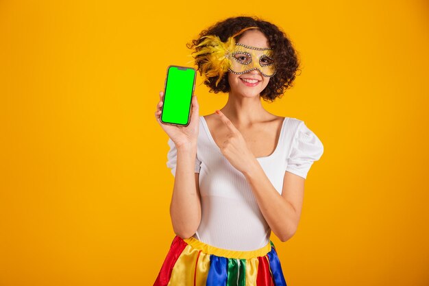 Beautiful Brazilian woman dressed in carnival clothes colorful skirt and white shirt holding smartphone and showing green screen in chroma