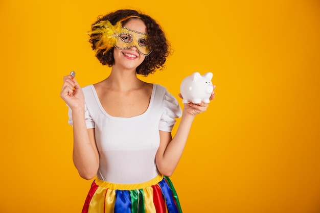 Beautiful brazilian woman dressed in carnival clothes colorful skirt and white shirt wearing mascara holding piggy bank and coin