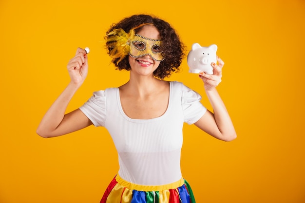 Beautiful Brazilian woman dressed in carnival clothes colorful skirt and white shirt wearing mascara holding piggy bank and coin