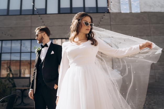 Beautiful bride in fashionable sunglasses and white dress stands in front of a handsome groom