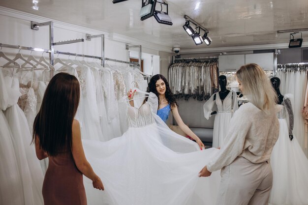 La bella sposa sta provando un abito da sposa elegante nel moderno salone di nozze che i suoi amici stanno aiutando