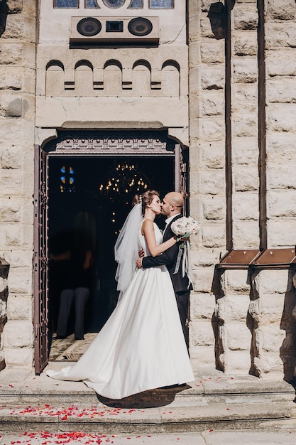 Beautiful bride kissing with stylish groom standing at church after wedding matrimony Gorgeous wedding couple after wedding holy ceremony Newlyweds