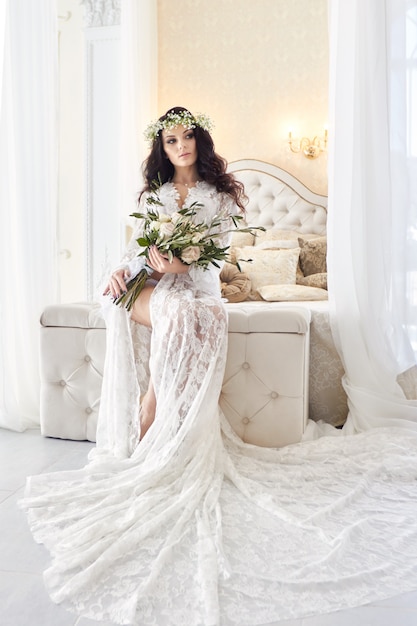 Beautiful bride in lingerie and with a wreath of flowers on her head, in the morning before the wedding. White negligee of the bride, preparing for the wedding ceremony. Sexy girl on the bed