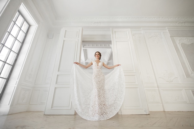 Bella sposa in posa in abito da sposa in uno studio fotografico bianco. specchio. divano. mazzo. la porta. lampadario.