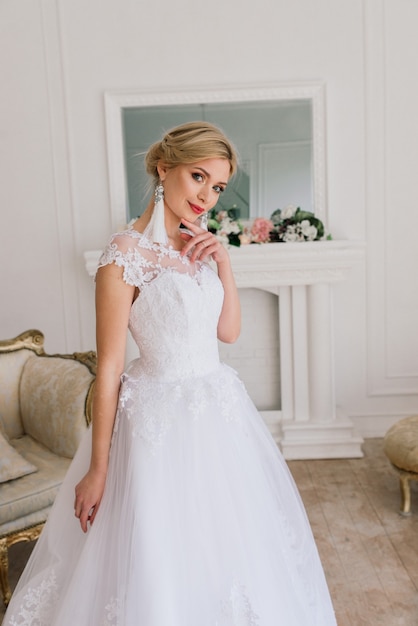 Beautiful bride posing in wedding dress in a white photo studio, mirror and sofa.