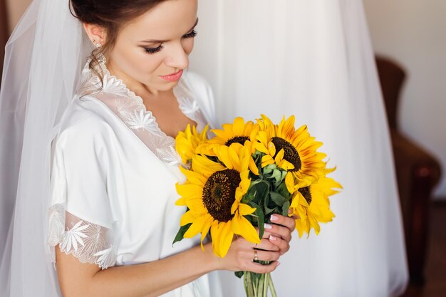 結婚式の朝にベールと花の緑豊かな花束とドレスの近くのローブの美しい花嫁