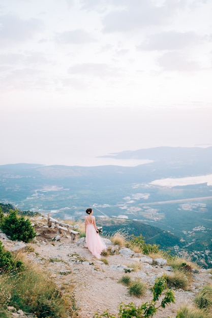 Foto bella sposa e in piedi sulla montagna, tenendo il bouquet e guardando la baia di kotor.
