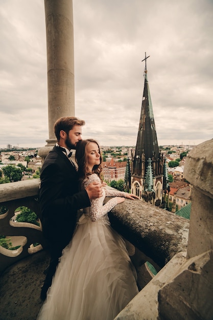 Photo beautiful bride and stylish groom are hugging on the balcony of old gothic cathedral with panoramic city views