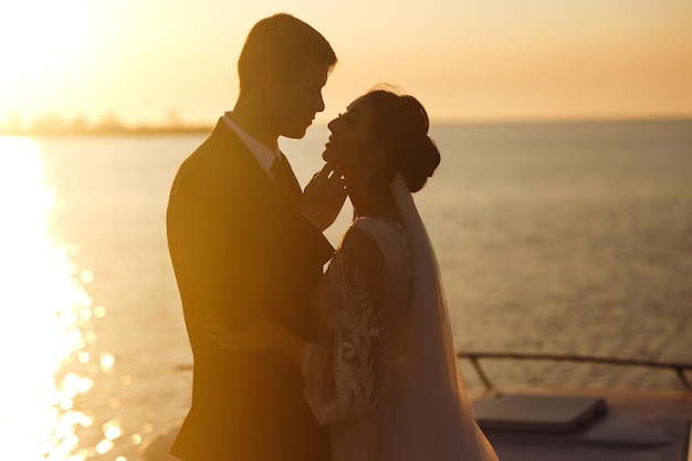 Beautiful bride and stylish groom together on the bridge against the background of boat at sunset