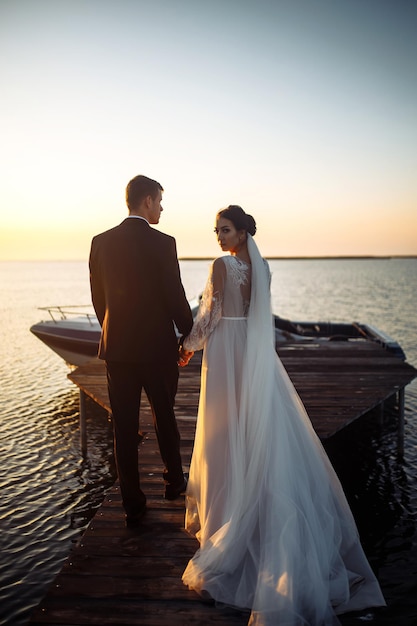 Красивая невеста и стильный жених вместе на мосту на фоне лодки на закате