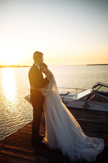 Beautiful bride and stylish groom together on bridge against the background of the boat at sunset