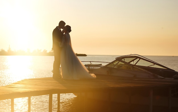 Beautiful bride and stylish groom together on the bridge against the background of boat at sunset