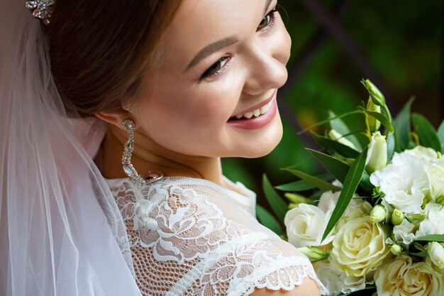 Beautiful bride in veil and white coat sits on a blanket with a wedding bouquet in her hands