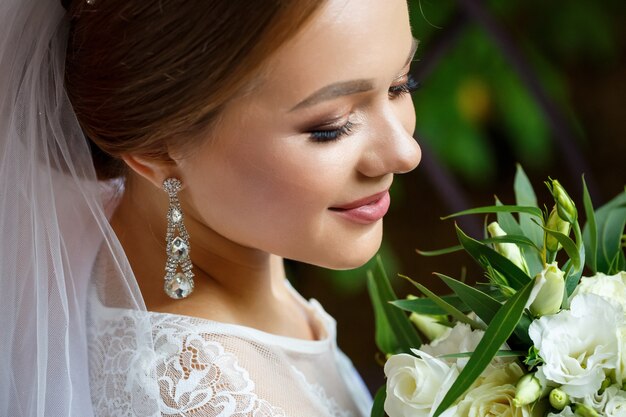 Beautiful bride in veil and white coat sits on a blanket with a wedding bouquet in her hands