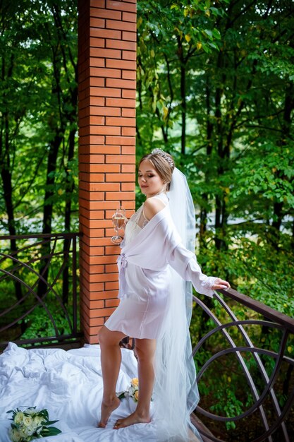 Beautiful bride in a white coat on the balcony drinks champagne from a glass