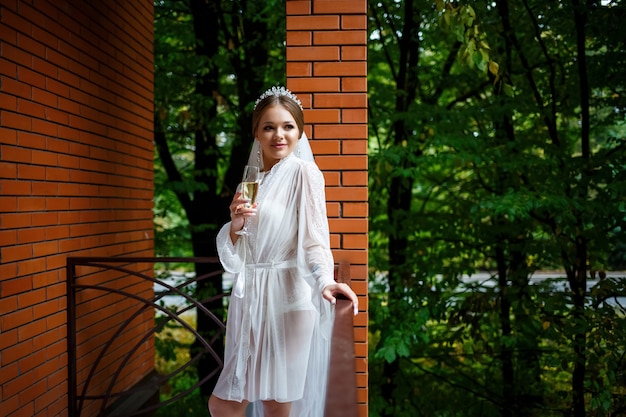 Beautiful bride in a white coat on the balcony drinks champagne from a glass