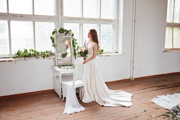 A beautiful bride in a white dress is sitting on a chair near the window and holding a wedding bouquet