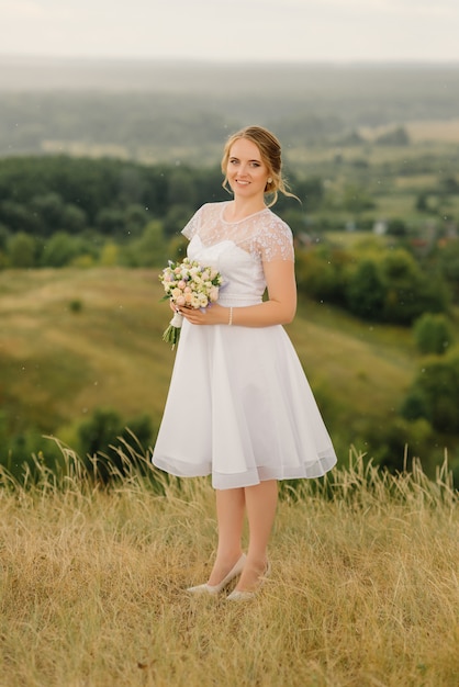 A beautiful bride in a white dress stands in nature and holds a wedding bouquet in her hands.