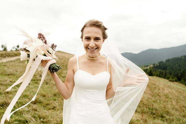 A beautiful bride in a white dress and with a wedding bouquet of dried flowers is in the autumn mountains among the yellow grass