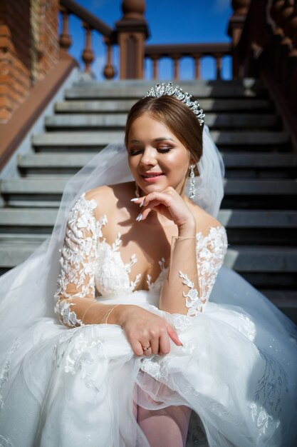 Beautiful bride in a white wedding dress sits on the steps and smiles beautifully