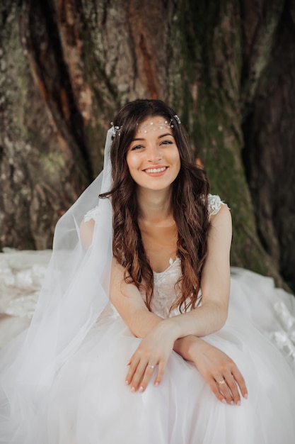 Фото Красивая невеста с длинными кудрявыми волосами в шикарном платье улыбается, глядя в линзу под большим деревом портрет невесты весенняя свадьба естественный макияж