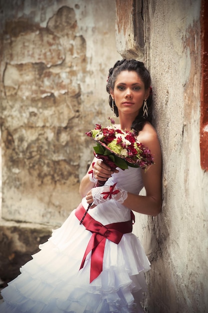 Beautiful bride with wedding bouquet in her hands leaning against the wall of an old building