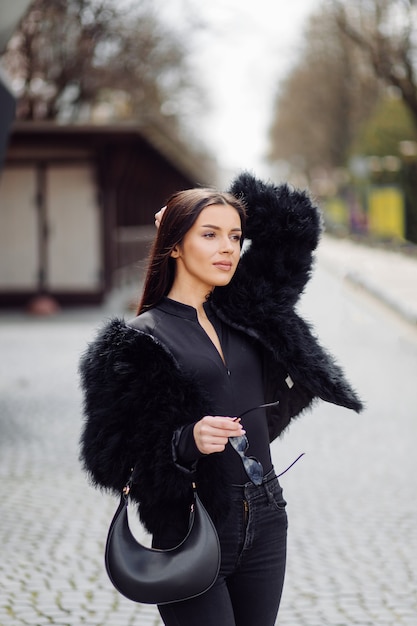 Beautiful brown-haired stylish girl in black dress outdoor. Young attractive elegant woman portrait with long hair on springtime in city streets.