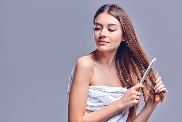 A beautiful brown-haired woman with bared shoulders looks at the camera as she brushes her hair with a wooden brush, isolated on a gray background with copy space for text.