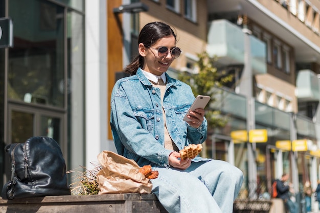 Bella bruna 20-30 anni che si gode cibo delizioso dal ristorante da asporto in strada all'ora di pranzo usando il telefono per valutare l'azienda di cibo da asporto per un menu delizioso